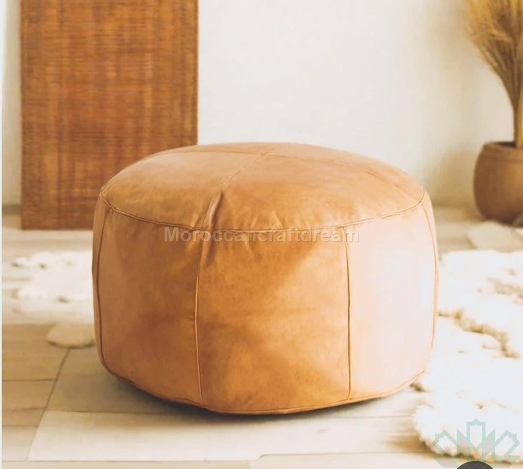 A large round brown leather coffee table pouf placed on a light-colored rug with a bamboo-like object in the background.