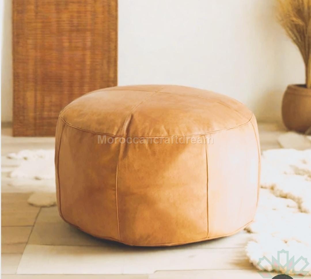 A large round brown leather coffee table pouf placed on a light-colored rug with a bamboo-like object in the background.